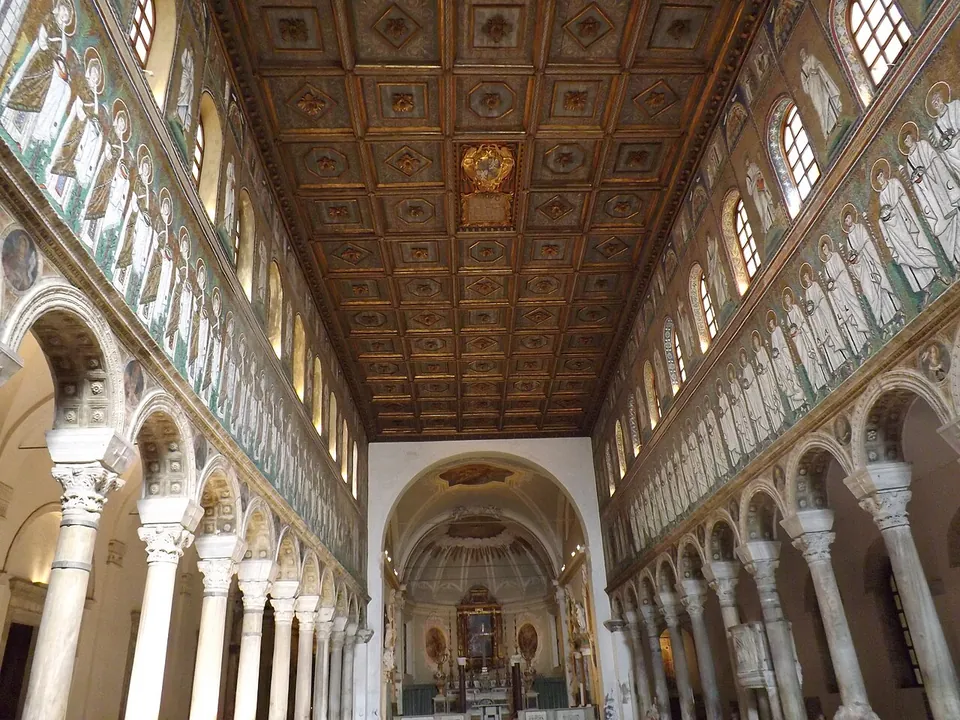 A long nave lined with arcades on white columns; above them, on both walls, a row of figures in white robes in mosaic on a gold ground, under a coffered wooden ceiling, with the apse at the far end.