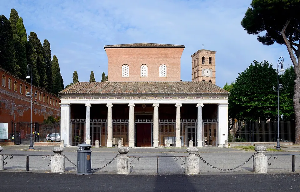 A plain brick church front with three arched windows above a long portico of six columns, a brick bell tower with a clock to the right, cypresses and umbrella pines around it and an empty paved square in front.