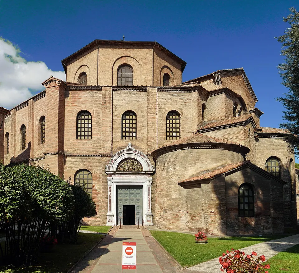 A tall brick church rising in stepped, many-sided storeys with round-arched windows, a white stone doorway at its base, seen across a lawn with a paved path and red flowers.