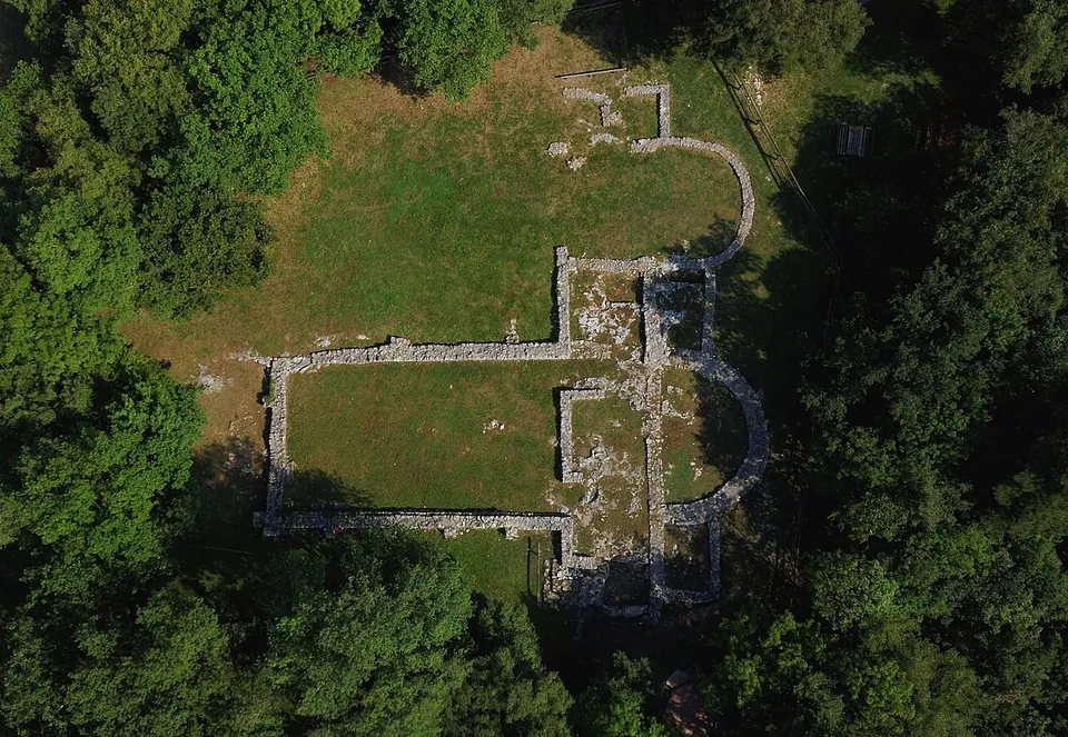An aerial view of low stone wall footings outlining a long rectangular hall and a smaller one beside it, each ending in a rounded apse, on grass in a clearing surrounded by trees.
