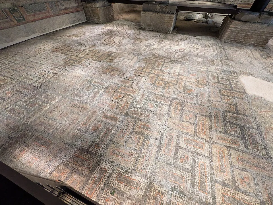 A wide mosaic floor of interlocking geometric patterns in muted red, grey and cream, seen in a dim underground room with brick piers and a low ceiling.