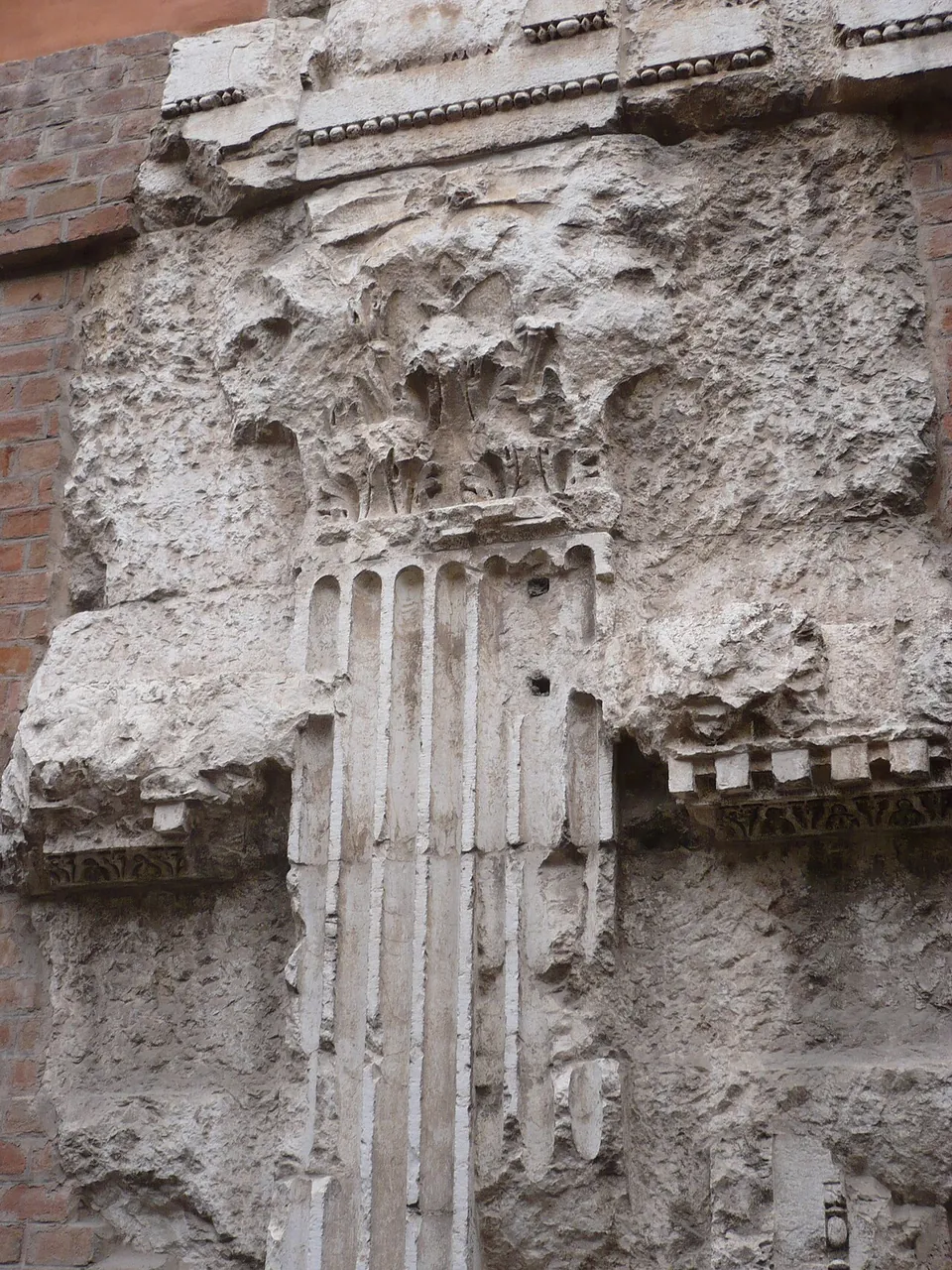 A weathered Corinthian pilaster capital of pale stone, the fluted pilaster below it and a band of carved moulding, set in a wall of rough masonry beside a strip of red brick.