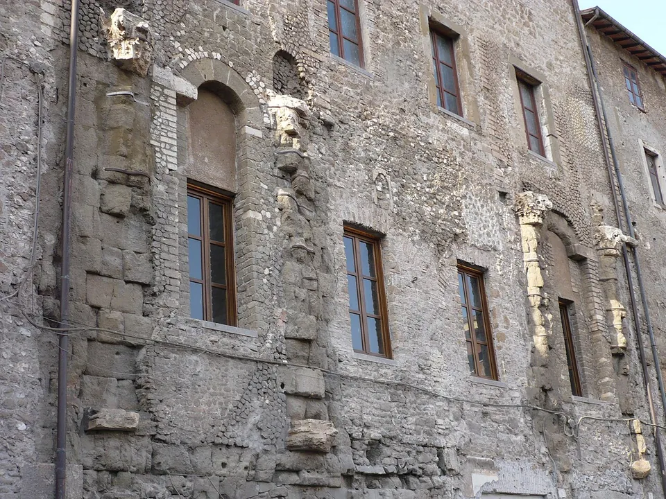 A rough stone wall with rectangular windows, between which the weathered shafts of ancient half-columns, carved capitals and the outline of an arch are set into the masonry.