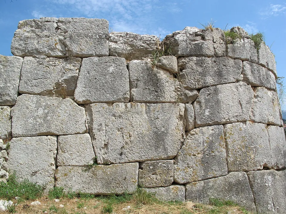 Close view of a curving face of massive limestone blocks, each cut to a different shape and fitted tightly against its neighbours without mortar, with tufts of grass growing from the top course.