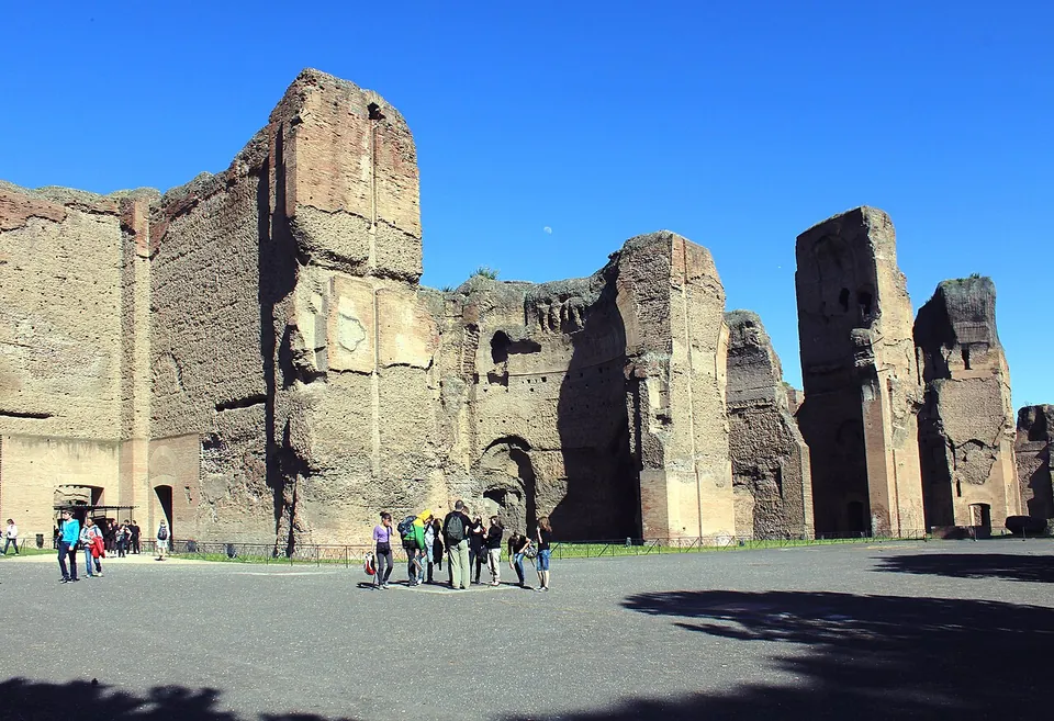 Massive brick walls and free-standing piers, several storeys high, of rough brick, standing over a wide open gravel area where a group of visitors is walking.