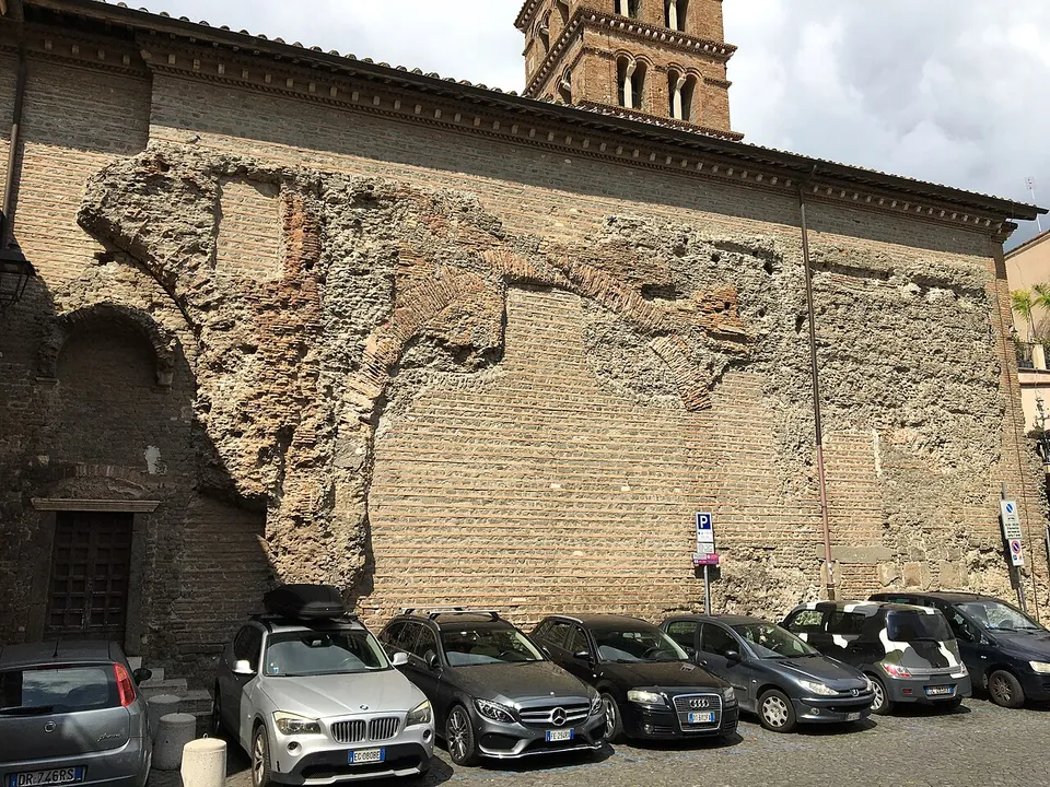 The long side wall of a church, its brick face broken away in places to show a rough core of stone and mortar, with a brick bell tower above and cars parked along its foot.