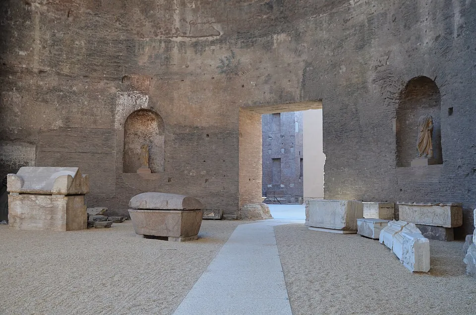 A lofty hall of bare brick with round-headed niches high in its walls, one holding a statue, and stone sarcophagi and carved blocks standing on a gravel floor either side of a paved path that leads to a doorway.