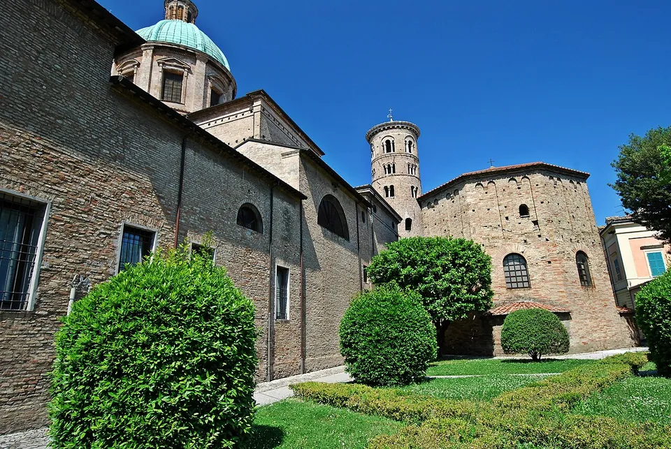 A plain octagonal brick building with a row of arched windows and a low tiled roof, standing in a garden of clipped hedges and lawn, between a long brick building with a green-domed church at its far end and a tall round brick bell tower behind.