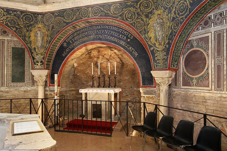 A deep arched niche with a small altar and candlesticks, framed by an arch of mosaic with a Latin inscription in white letters on dark blue, and beyond it walls panelled in coloured marble with a round stone inlay, behind a low railing and a few chairs.