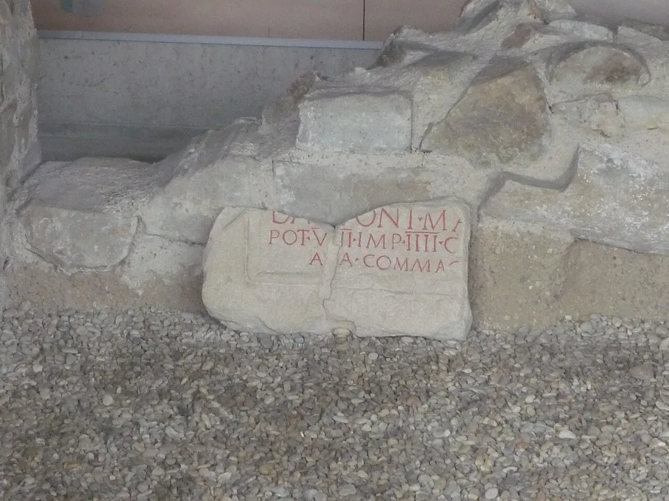 A squared block of pale stone set among rough masonry and gravel, with a few lines of Roman capital letters picked out in red.