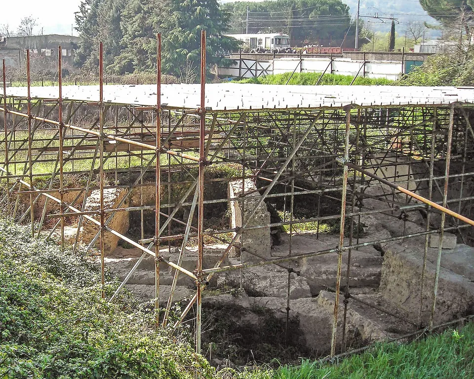 Excavated stone foundations in a hollow, covered by a frame of metal scaffolding poles and a sheet roof, with grass in front and a railway line and trees behind.