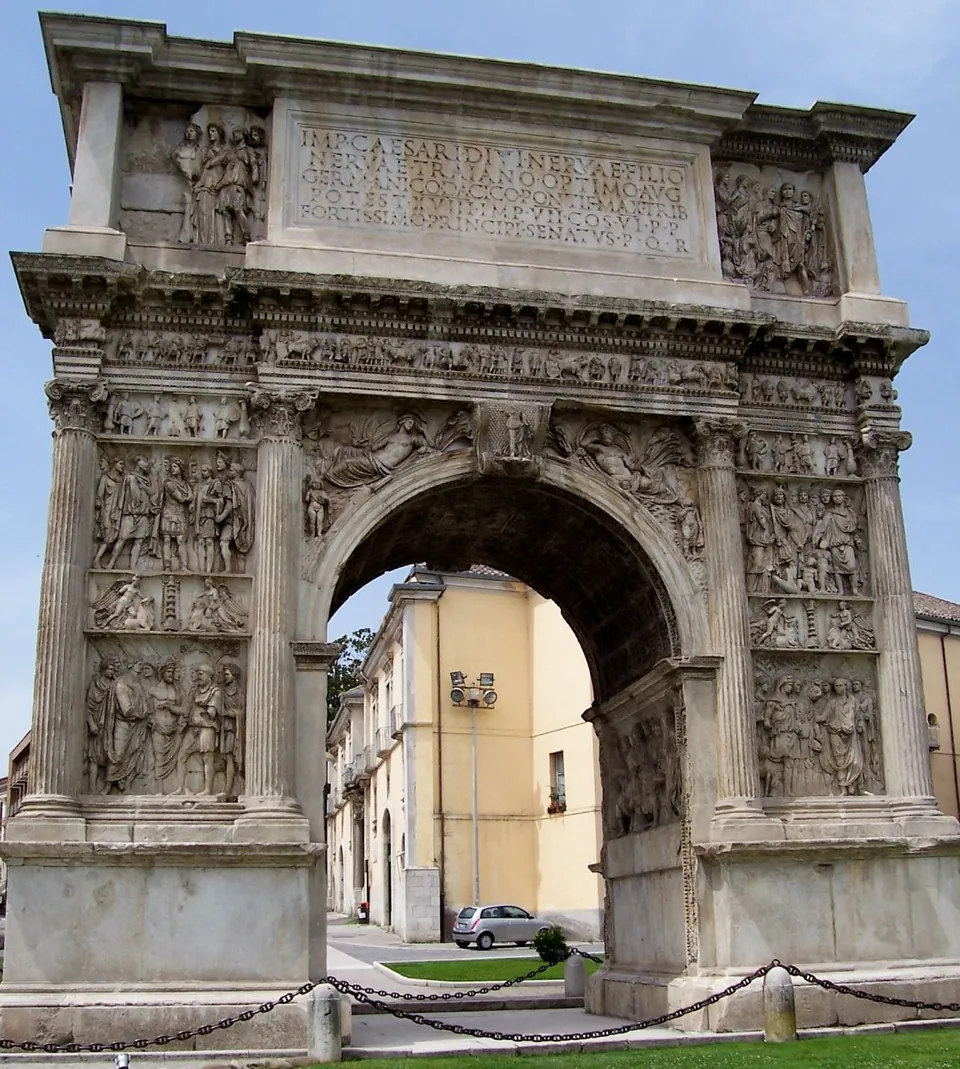 The arch seen straight on from the other side, its piers covered in relief panels between fluted columns and its attic carrying the same long inscription, with a pale house and a parked car visible through the archway.