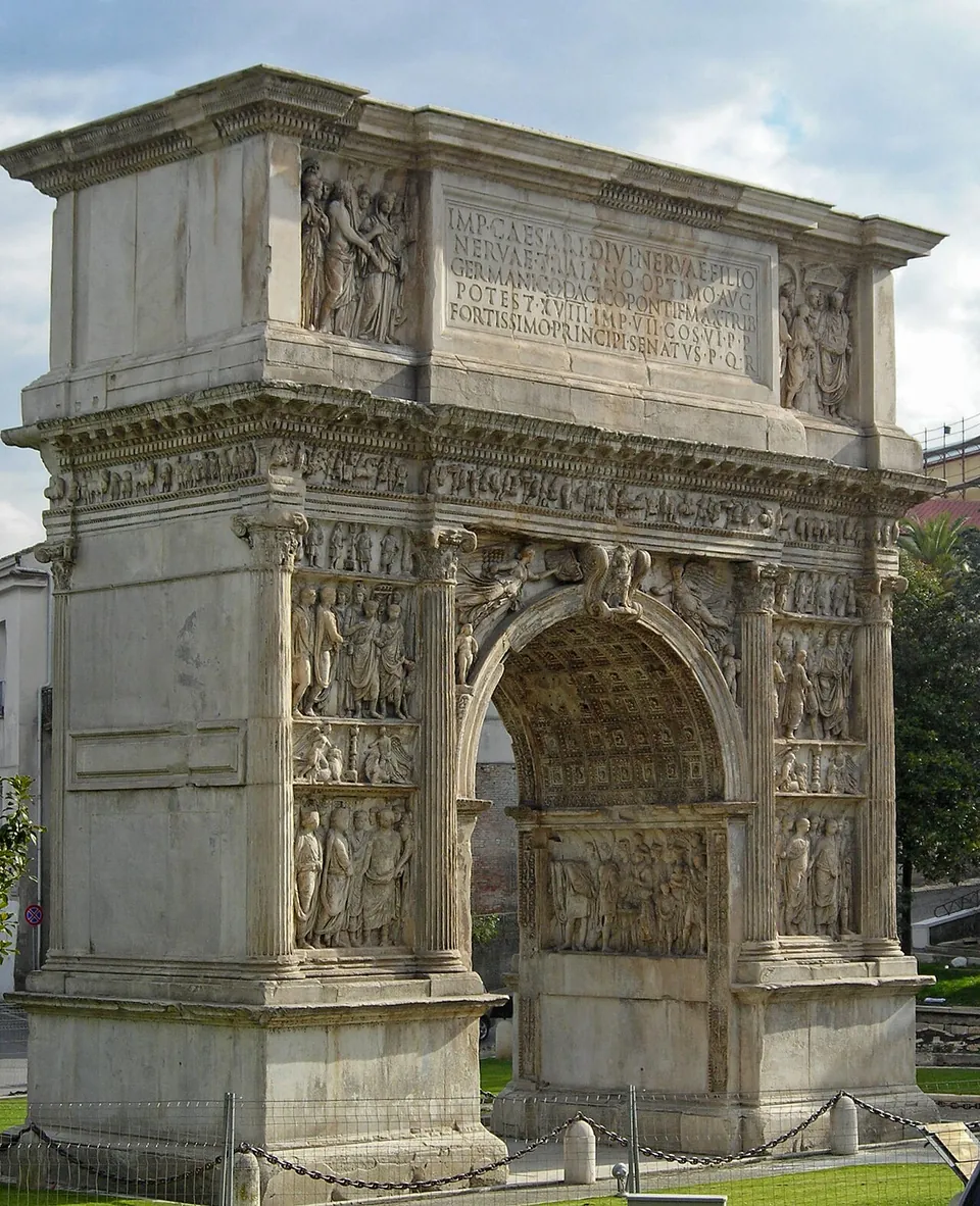 A single-arched marble gateway seen at an angle, every surface of its two piers carved with stacked relief panels of standing figures between engaged columns, a carved frieze running beneath an attic with a long Latin inscription flanked by two more relief panels, and a coffered vault visible inside the arch; a low chain barrier runs along its foot.