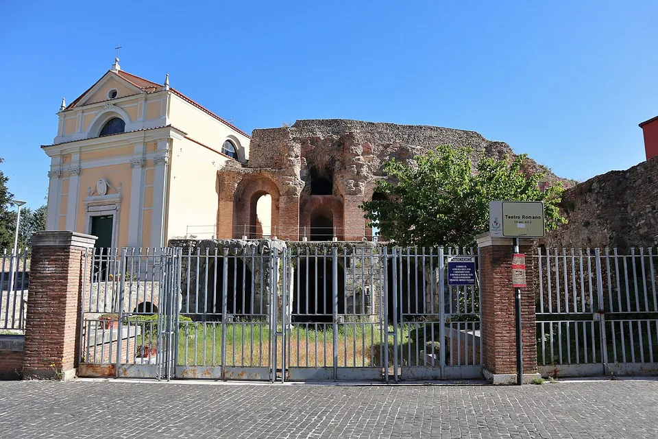 A gated entrance in a railing between brick pillars, with a pale-fronted church on the left and the ruined arches of the theatre's outer wall rising behind the fence.
