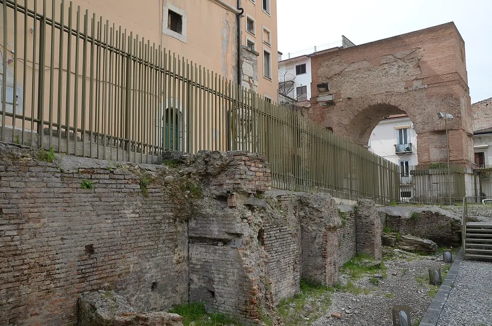 Low ruined walls of brick and rubble below a tall metal fence, with a gravel path and a short flight of steps beside them and the brick arch rising beyond.