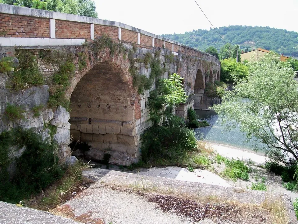 A low bridge of stone and brick arches crossing a shallow river, overgrown with plants at its foot, with a wooded hill behind.