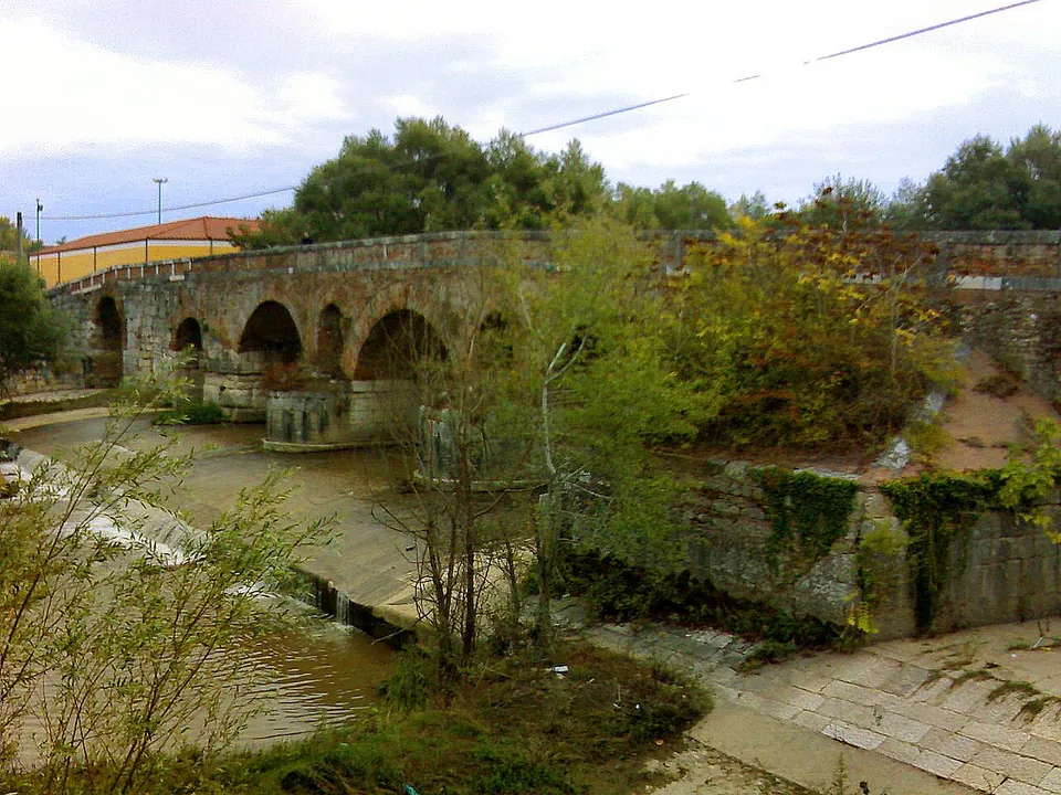 A long bridge of stone and brick arches crossing a shallow river under trees, with overgrown banks and a stone embankment in the foreground.
