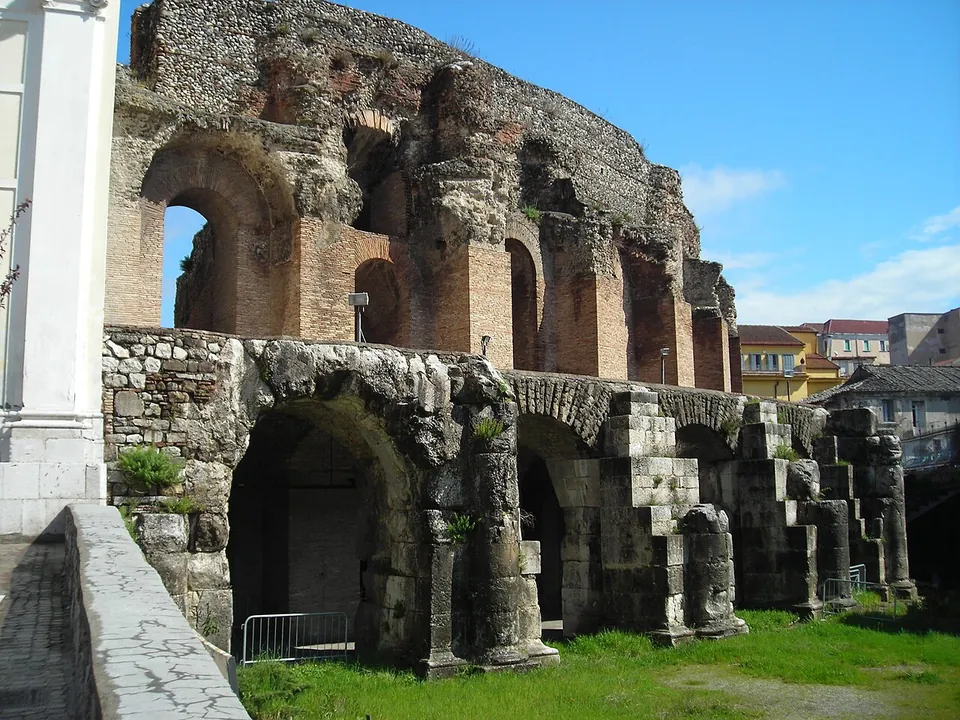 The outside of the curved theatre wall, a row of stone arches at ground level with half-columns against the piers, and above it a higher storey of brick-and-stone arches, broken off at the top.