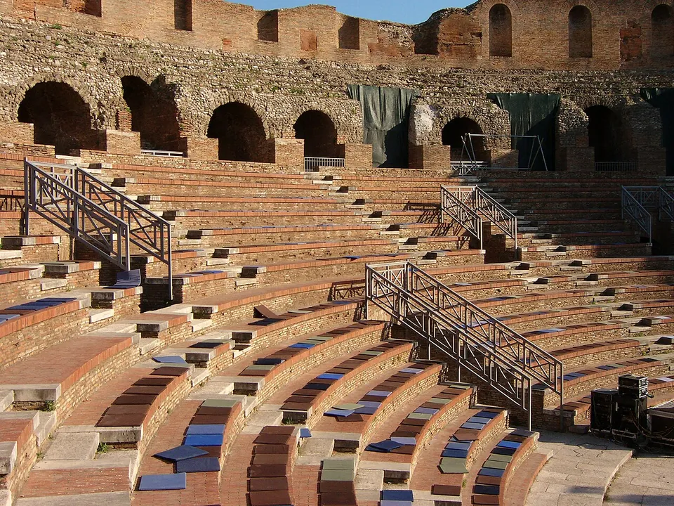 Curved rows of brick-and-stone seating climbing towards a ring of round-headed arches at the top, with metal stair railings running down between the rows and coloured pads laid along some of the steps.