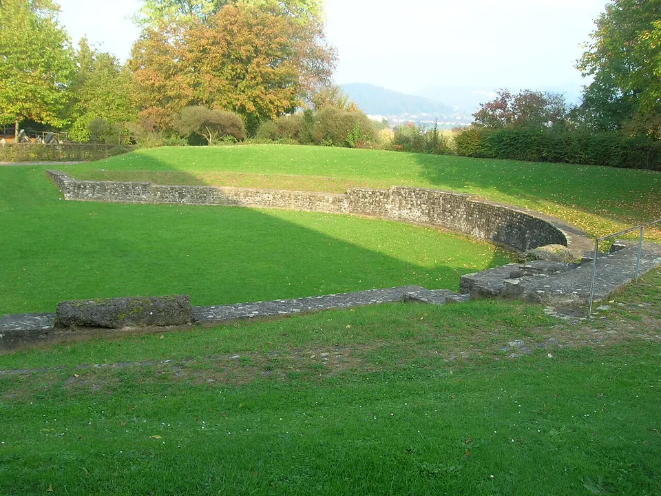A small oval arena of mown grass enclosed by a low curved stone wall with an entrance gap, set in parkland with autumn trees and hills on the horizon.