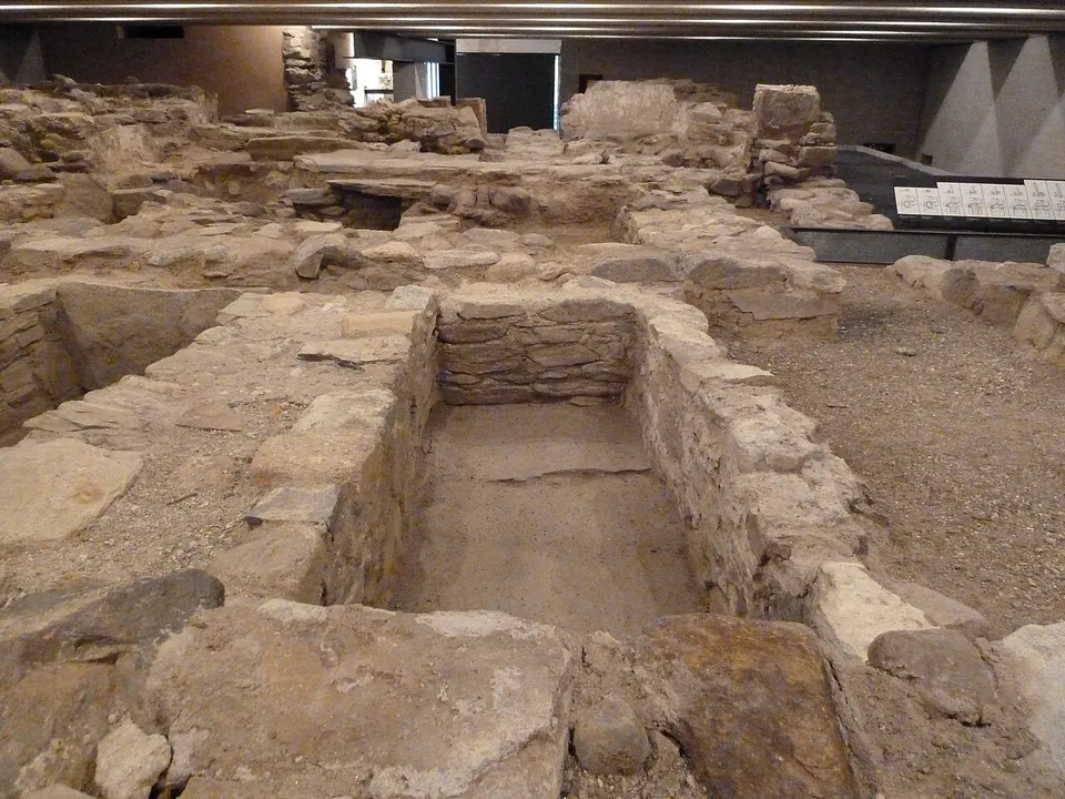Excavated walls of rough stone under a concrete roof, with a rectangular stone-lined grave in the foreground and information panels along a walkway at the back.