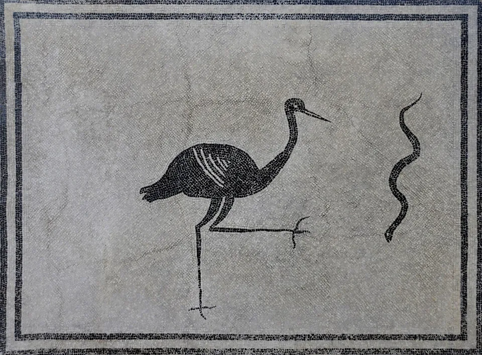 A black-and-white mosaic panel inside a double black border, showing a long-legged black bird striding with one foot raised towards a wriggling black snake.