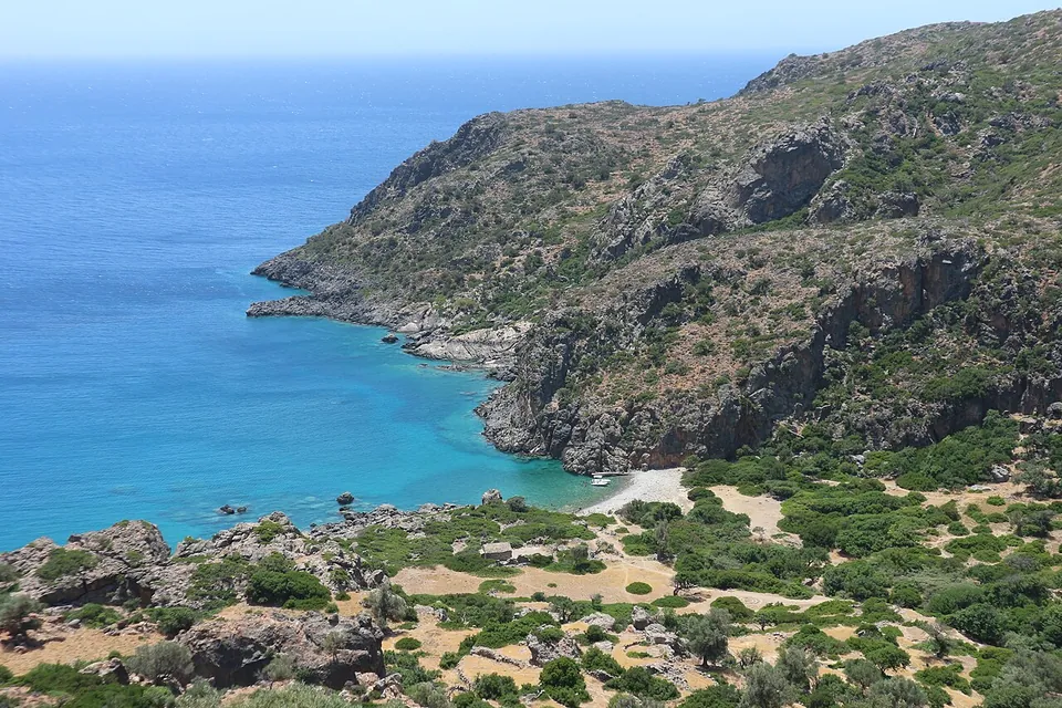 A small bay of turquoise water below steep rocky hills, with a pale pebble beach and a green valley of trees behind it.