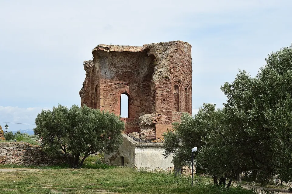 A tall ruined wall of red brick with round-arched windows and blind arcading, rising above olive trees and a lawn.