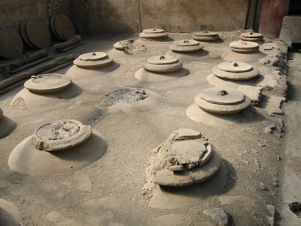 Rows of round lidded storage jars sunk to their necks in a dusty earth floor, with more lids stacked against the wall behind.