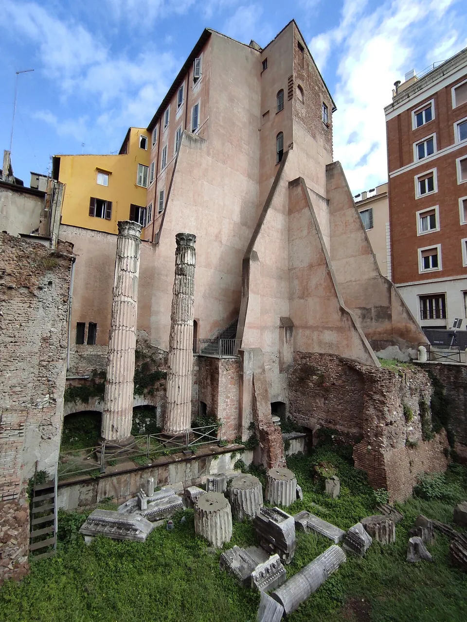 Two fluted column shafts standing in a sunken, grassy excavation, with column drums and carved blocks lying in front of them and brick walls at the side, below the tall, buttressed blank wall of a modern building.