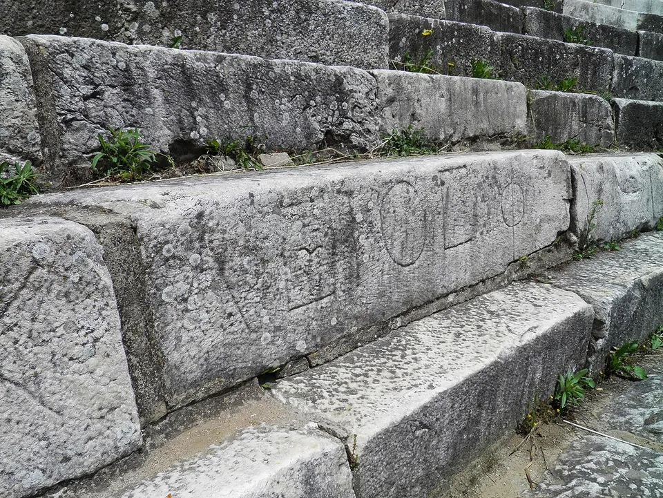 Worn grey stone seat blocks in stepped rows, the front of one carved with large Greek letters.