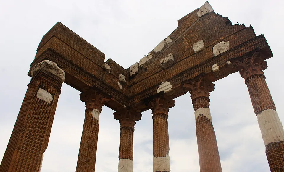 Looking up at the corner of a temple porch against a pale sky, Corinthian columns and the beam above them built of red brick with pieces of white stone set into them.