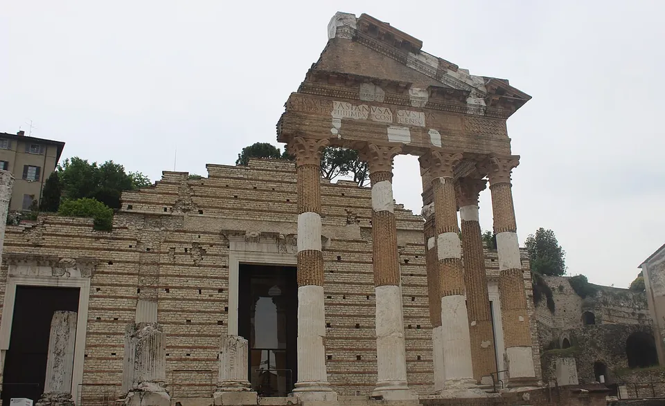 The front of a Roman temple seen straight on, a porch of columns banded in pale stone and red brick carrying a broken pediment with a fragment of carved inscription, and behind it a wall of pale squared stone with three dark doorways.