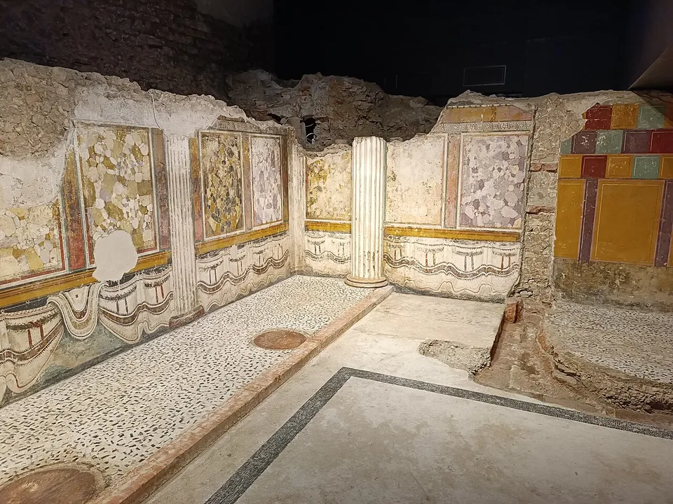 A dim, low room whose walls are painted with framed panels imitating coloured marble above a painted hanging curtain, a white fluted half-column standing at the far end and a floor of small pale stones with a dark border.