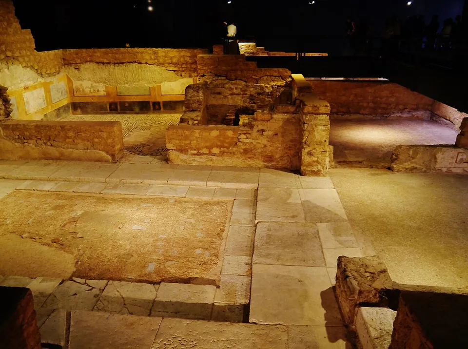 The low walls of excavated Roman rooms under a museum roof, lit warmly against the dark, with stone paving, a floor panel and fragments of painted plaster on the walls.