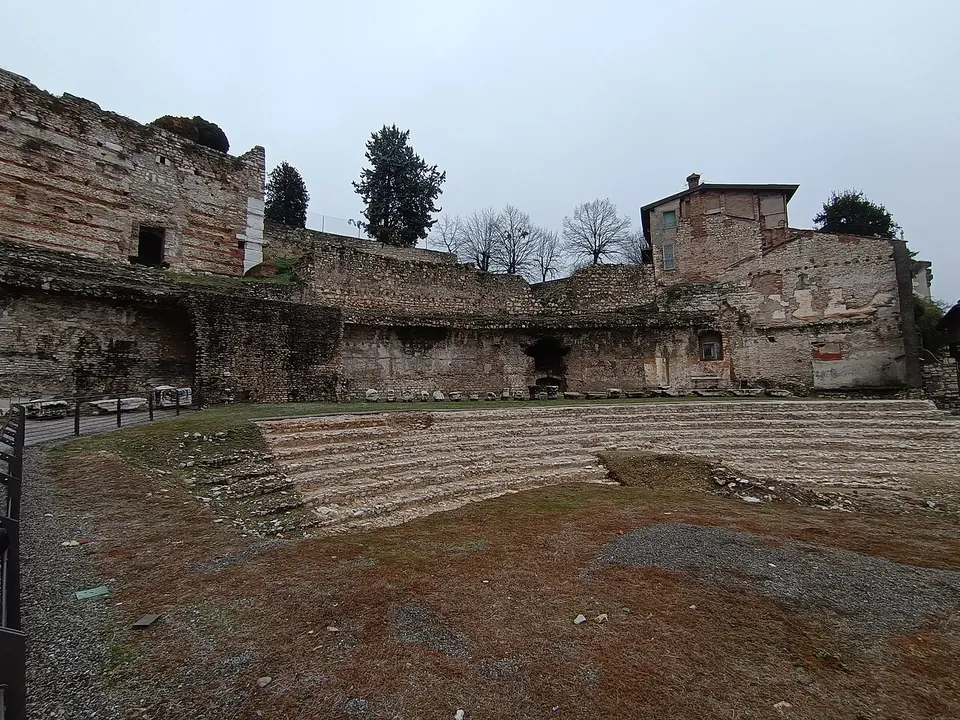 A curve of low stone seating tiers rising from bare ground, backed by tall ruined walls of stone and brick with openings in them and a brick house on top, under a grey sky.