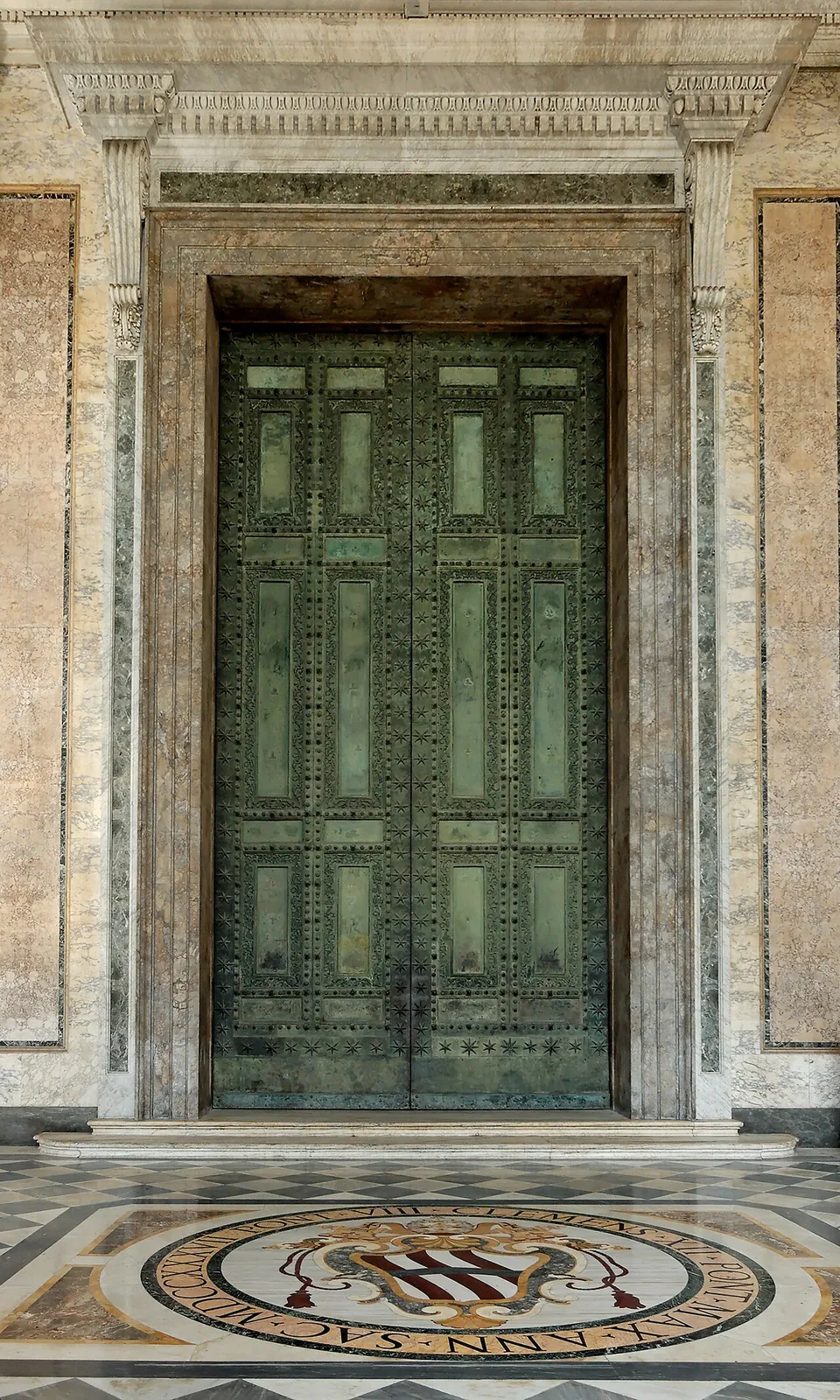 A tall double door of green-patinated bronze divided into rectangular panels studded with small bosses, set in a pale marble frame above a patterned marble floor.