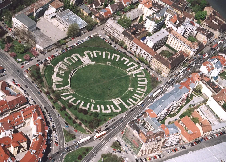 An aerial view of a large grassed oval enclosed by a ring of short radiating stone walls, set in a green lawn and surrounded on all sides by roads, parked cars and blocks of flats.