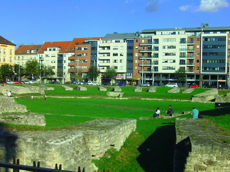 People sitting on the grass inside the arena on a sunny day, with low stone wall blocks around its edge and apartment buildings and parked cars beyond.