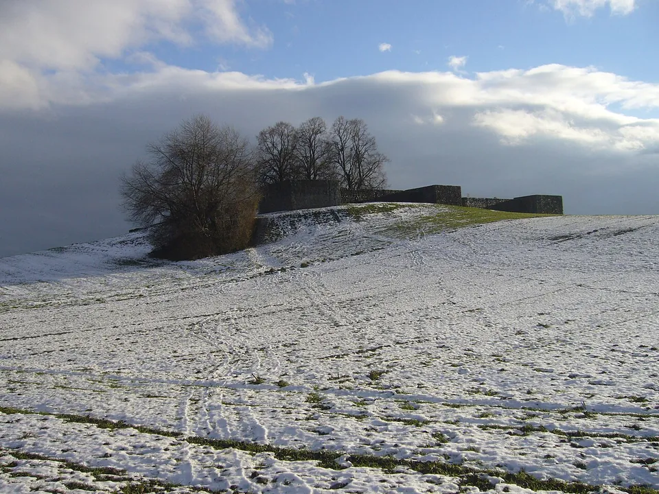 A snow-covered field rising to a low hilltop crowned by a line of stone walls and towers, with bare trees beside them under a cloudy sky.