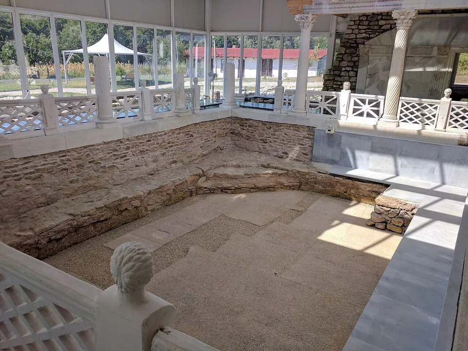 A sunken rectangular basin with stone sides and a sandy floor, surrounded by a white openwork marble balustrade topped by a carved head, with re-erected columns and the glass walls of the modern shelter behind.
