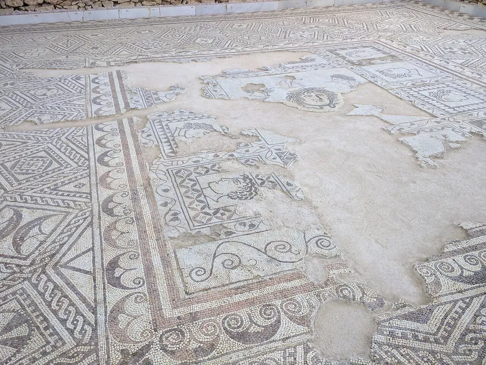 A close view of a worn mosaic floor in black and white tesserae, with bands of scrollwork and geometric borders round a damaged central field in which a face looks out of a small roundel.