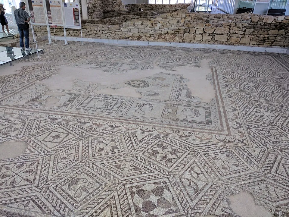 A large floor mosaic of interlocking geometric panels in black, white and ochre filling a room under a modern shelter, with low stone walls on two sides, information boards along the back and two visitors at the edge.