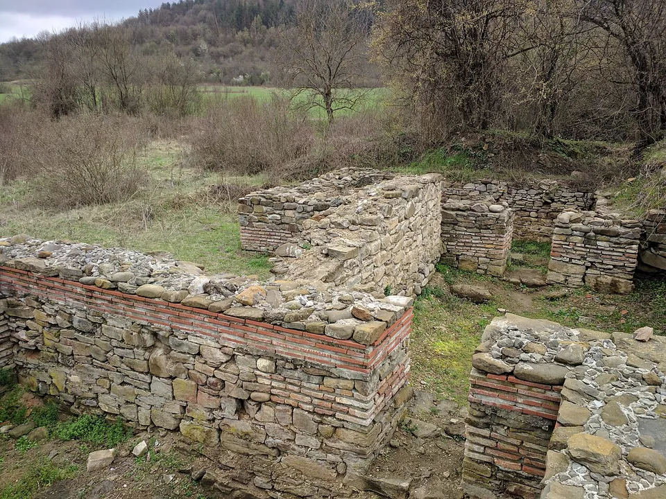 Close view of restored walls of flat stones laid in courses with bands of red brick, forming small rooms, with bare trees and fields behind.