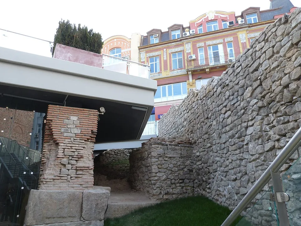 A square pier of alternating brick courses and stone blocks standing beside a tall wall of rough stone, below a modern concrete walkway, with apartment buildings above.
