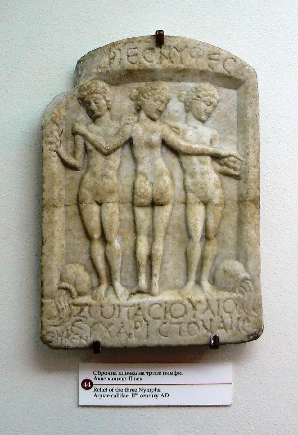 A marble relief of three standing nude female figures with their arms on each other's shoulders, between two water jars, with Greek letters above and below, hanging on a museum wall above a printed label.