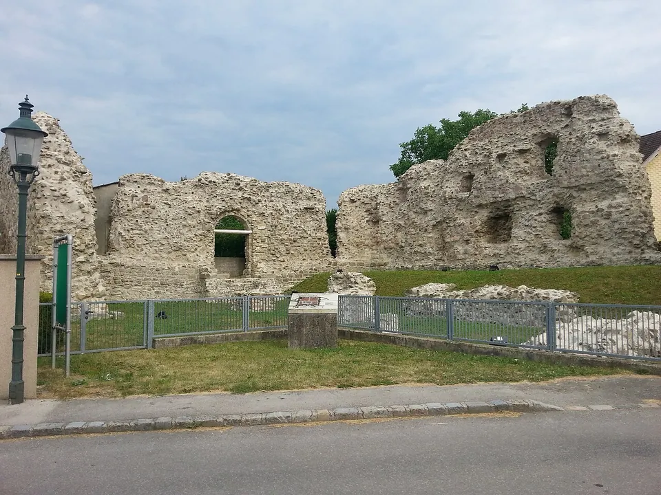 Tall broken walls of rough rubble masonry with an arched opening, standing in a fenced grassy plot behind a low railing and an information panel, beside a village street with a lamp post.