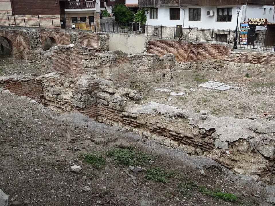 Excavated walls of stone with courses of brick enclosing several rooms, below street level, with a railing and modern houses behind.
