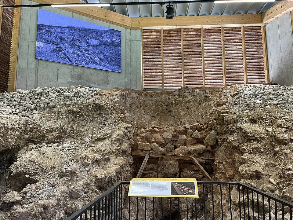 A deep, roughly round pit lined with rounded stones and gravel under a modern timber shelter, seen from a railed walkway, with an information panel at the front and a projected image on the far wall.