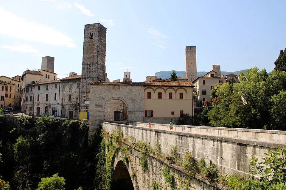 A stone bridge with a solid parapet, its arch half hidden by ivy and trees, crosses a wooded gorge towards a stone gateway, behind which rise tall square stone towers and the houses of the old town under a blue sky.